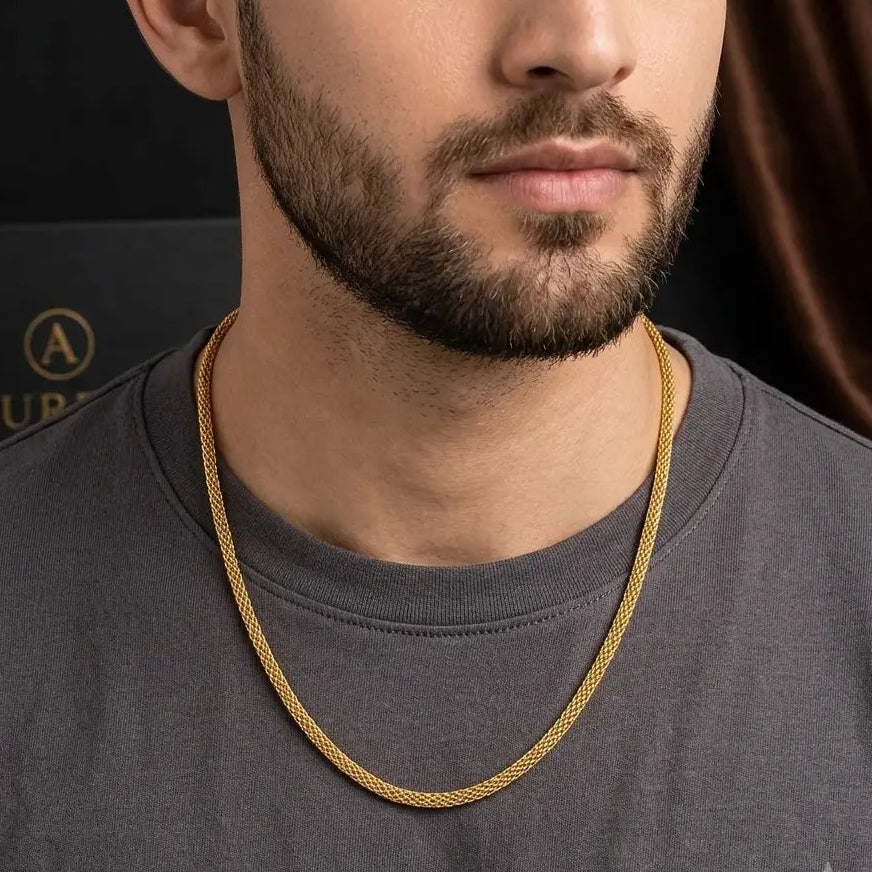 A close-up of a man wearing a gold chain necklace over a dark gray t-shirt, with a black branded box partially visible in the background.
