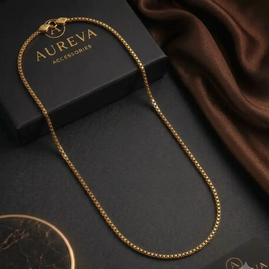 A gold chain necklace with a lobster clasp is displayed on a dark textured surface next to its black branded box, a brown satin fabric, and a round marble-like accessory.