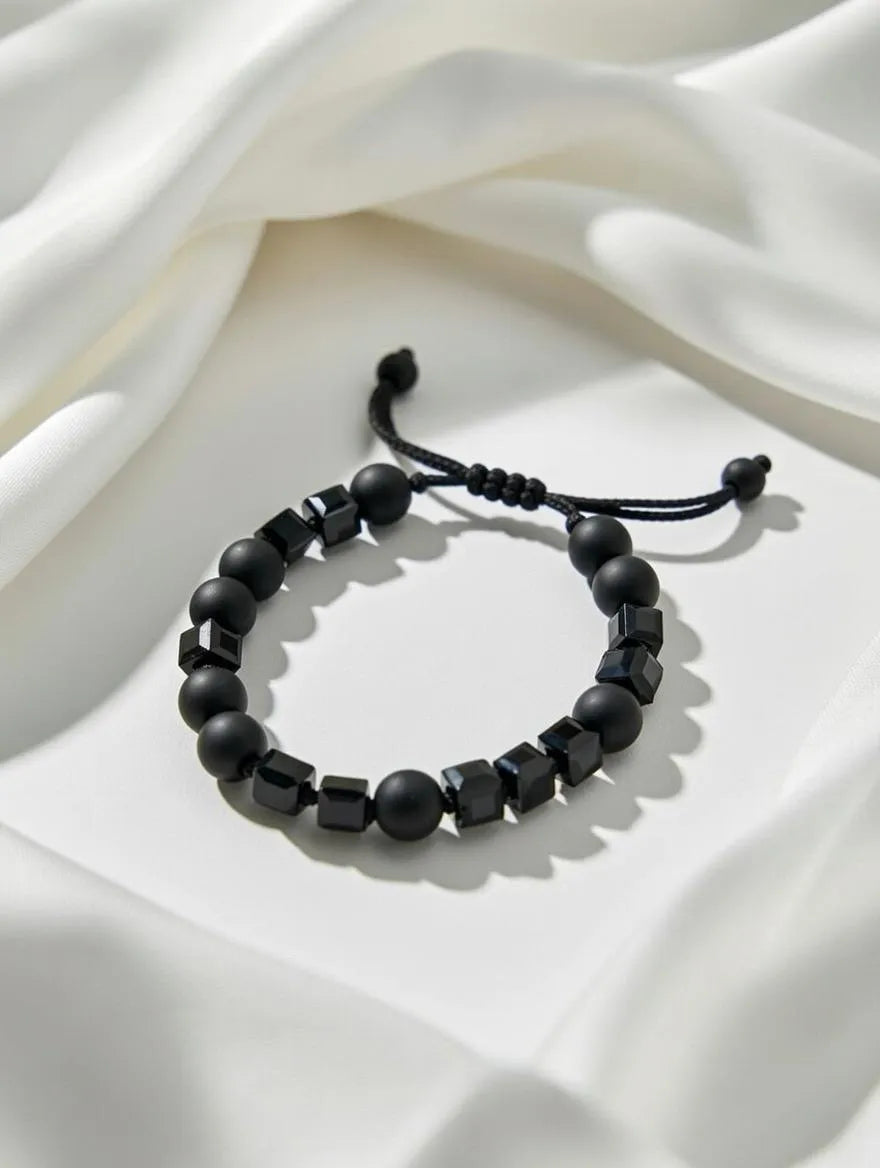 Handmade men's bracelet featuring matte black round onyx beads and polished black geometric cube beads on an adjustable black drawstring cord, displayed on a white silk background.