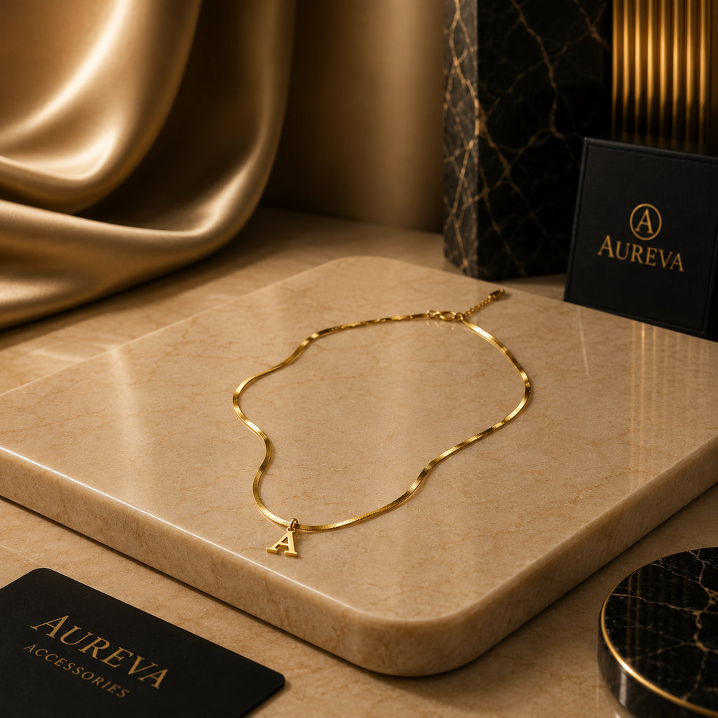 A gold wavy chain necklace with a small letter 'A' pendant is displayed on a beige stone slab, with Aureva accessories branding cards and a black marble box in the background.