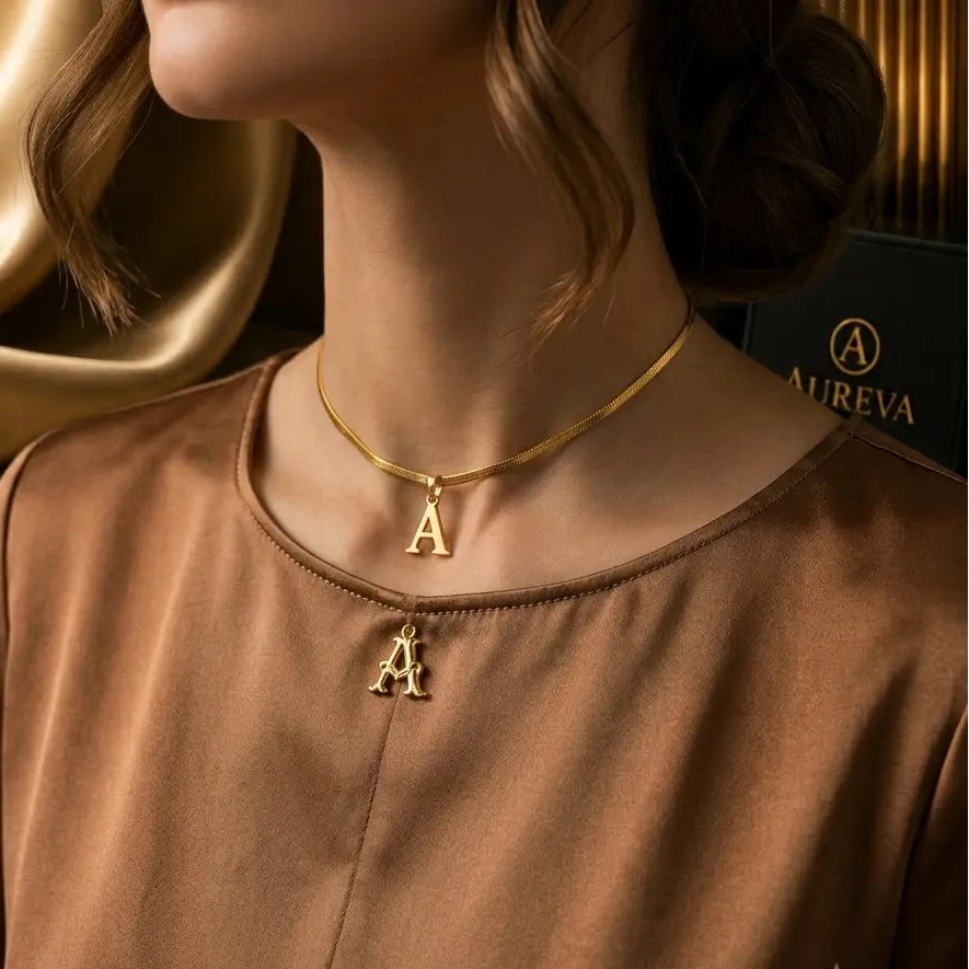 A close-up of a woman wearing a gold chain necklace with two hanging letter 'A' pendants, styled with a brown satin top and luxury jewelry packaging in the background.