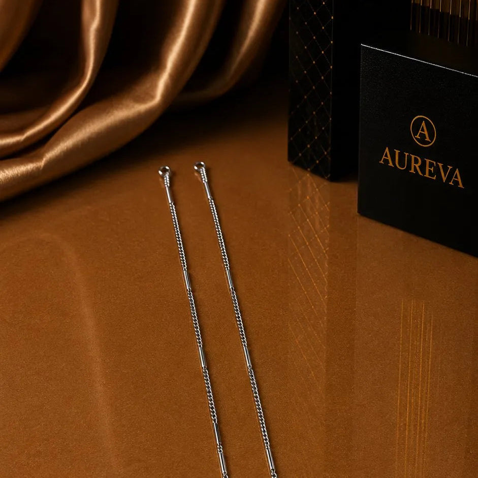 Two slim silver chain necklaces displayed on a brown presentation tray, with Aureva brand packaging and draped gold fabric in the background.