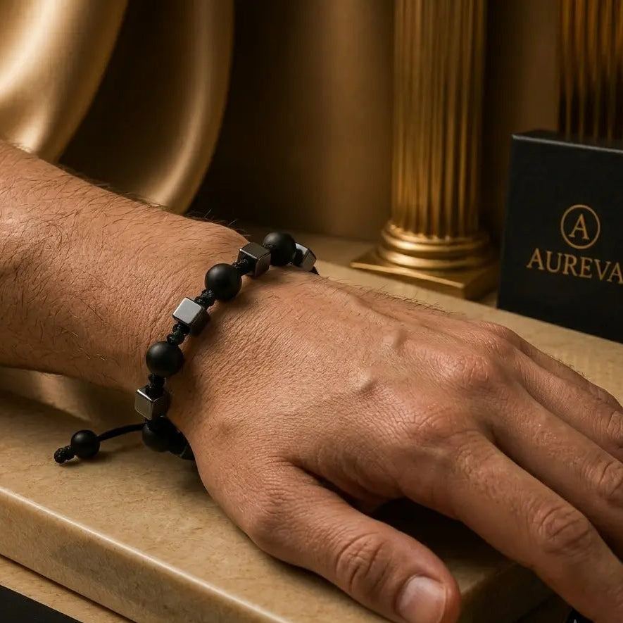 A close-up of a person's hand wearing a black beaded bracelet with metallic square accents, resting on a beige stone surface with Aureva branding visible in the scene.
