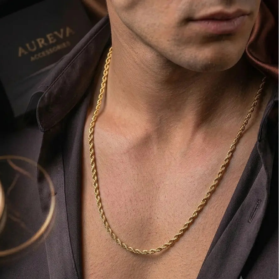 A close-up lifestyle shot of a man wearing a gold chain necklace with a twisted rope design, styled with a dark satin shirt and a black Aureva Accessories box in the background.