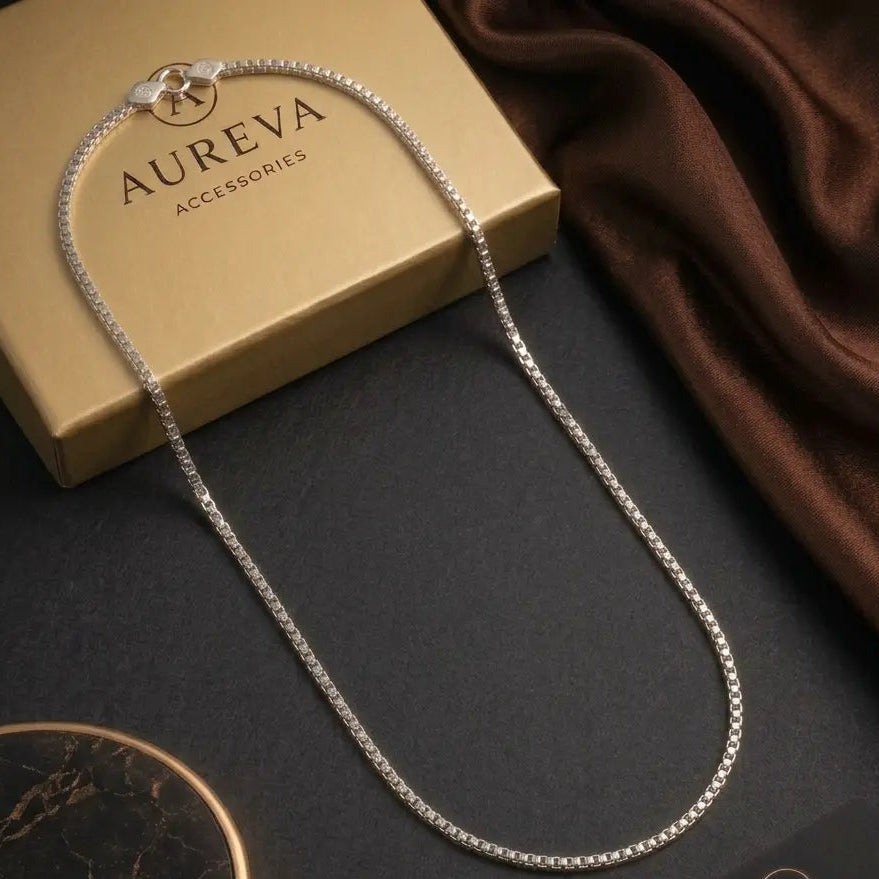 A silver chain necklace with a small gold-toned charm, draped over a gold Aureva Accessories box on a dark surface with a brown satin cloth and a branded card.