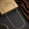 A silver chain necklace with a small gold-toned charm, draped over a gold Aureva Accessories box on a dark surface with a brown satin cloth and a branded card.