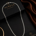 A silver-toned curb chain necklace draped over black Aureva Accessories boxes with a brown satin fabric in the background.