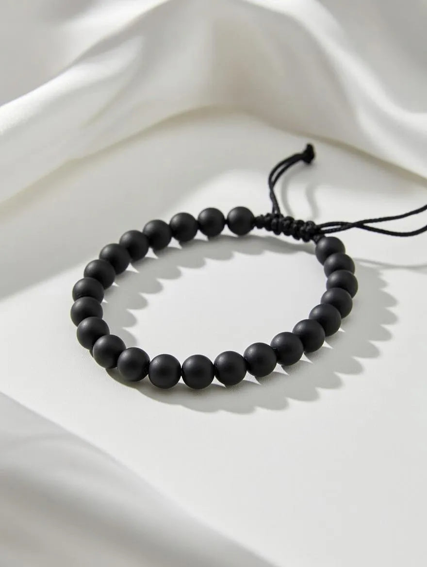 Unisex matte black onyx beaded bracelet with an adjustable black cord closure displayed on a soft white fabric background.