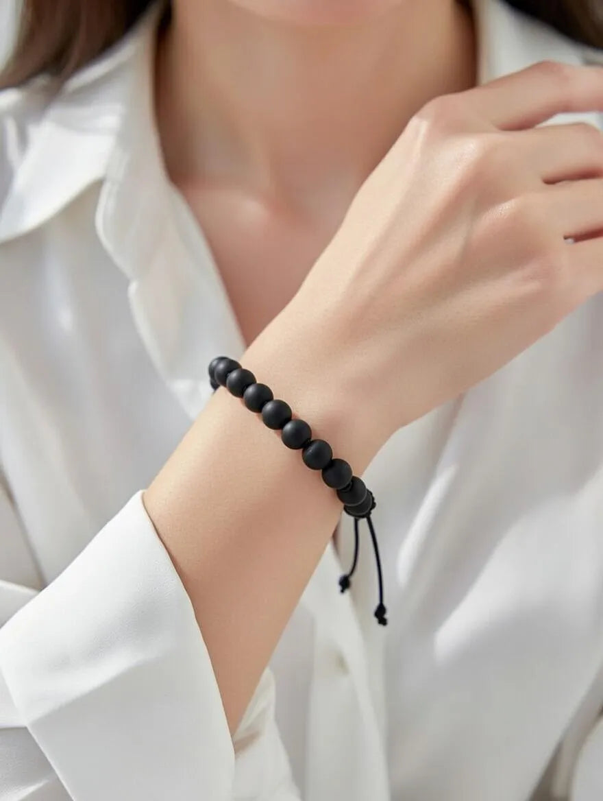 Unisex matte black onyx beaded bracelet with an adjustable black cord closure displayed on a soft white fabric background.