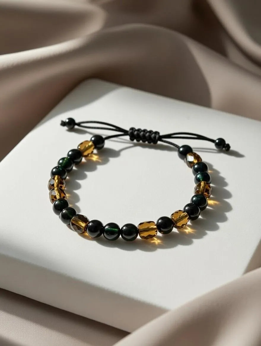 Unisex adjustable bracelet with green obsidian beads and faceted amber-colored tiger eye beads on a black cord, displayed on a white podium with silk fabric.