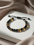 Unisex adjustable bracelet with green obsidian beads and faceted amber-colored tiger eye beads on a black cord, displayed on a white podium with silk fabric.