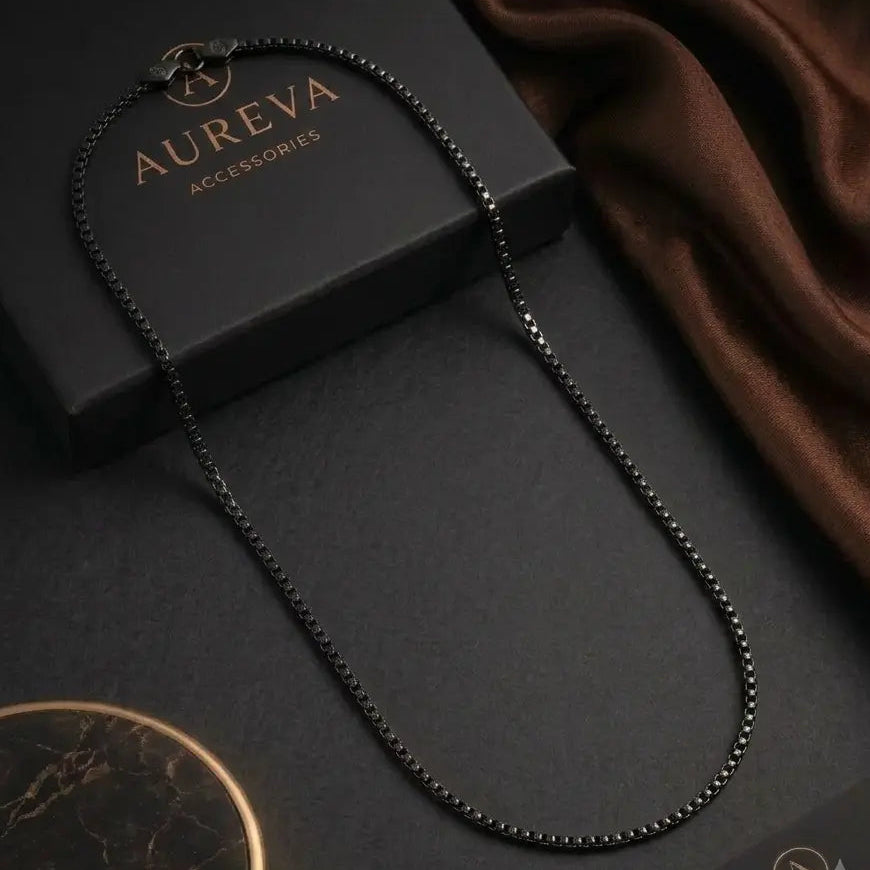 A black beaded necklace displayed on a dark surface with branded black boxes and a brown satin fabric. The necklace has a small circular pendant at the clasp.