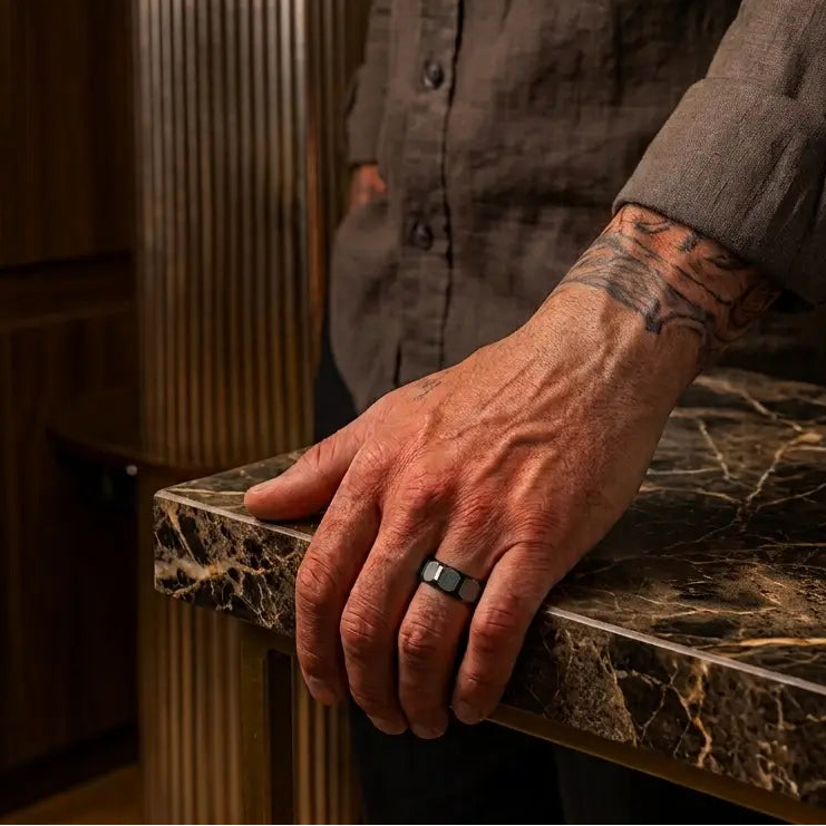 A close-up of a man's tattooed hand resting on a dark marble countertop, wearing a silver ring with a black inlay on his finger. He is wearing a dark brown linen shirt in a warm, modern interior setting.