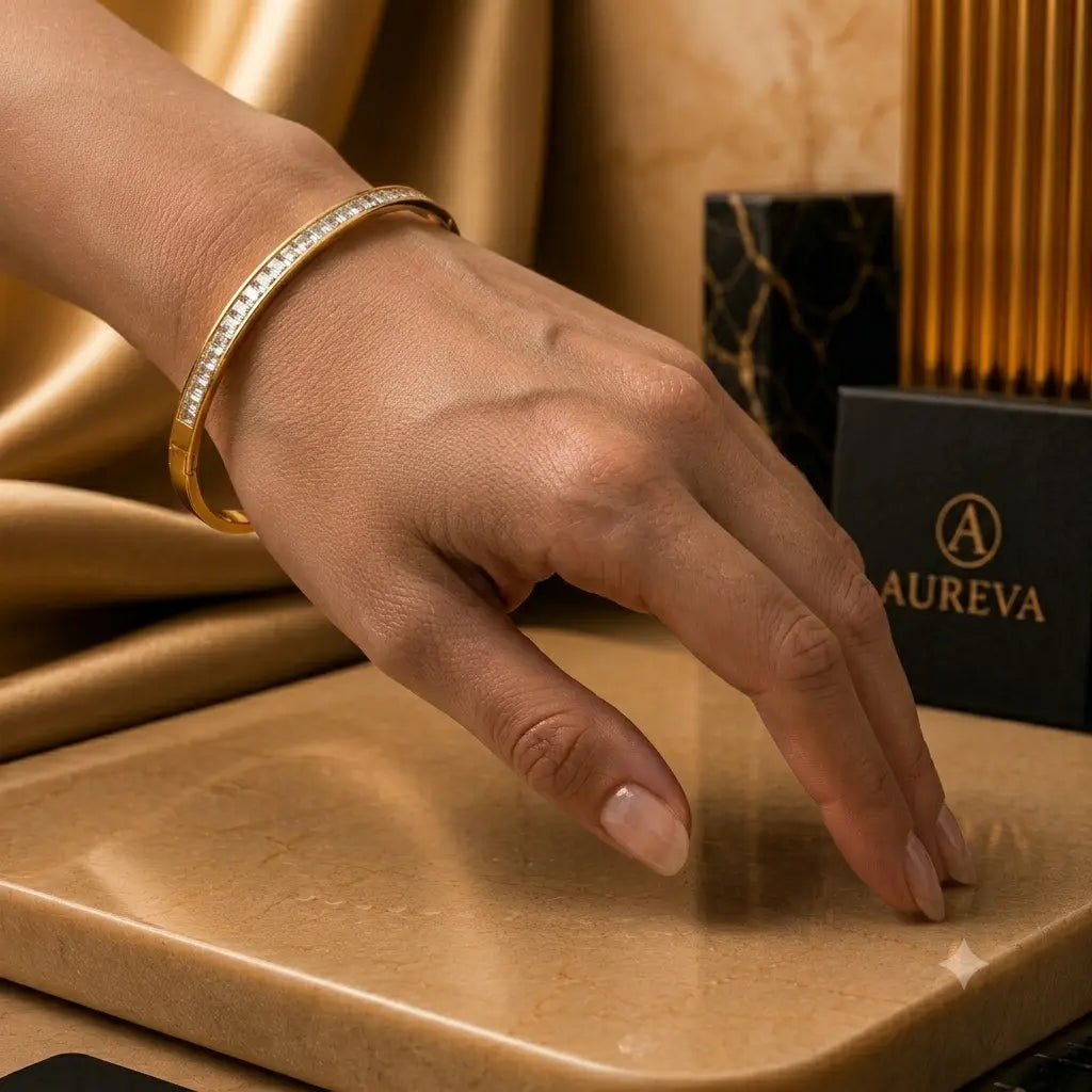 A close-up of a hand wearing a gold bangle bracelet with a row of clear stones, resting on a beige stone surface with a gold fabric backdrop and a black AUREVA box in the background.