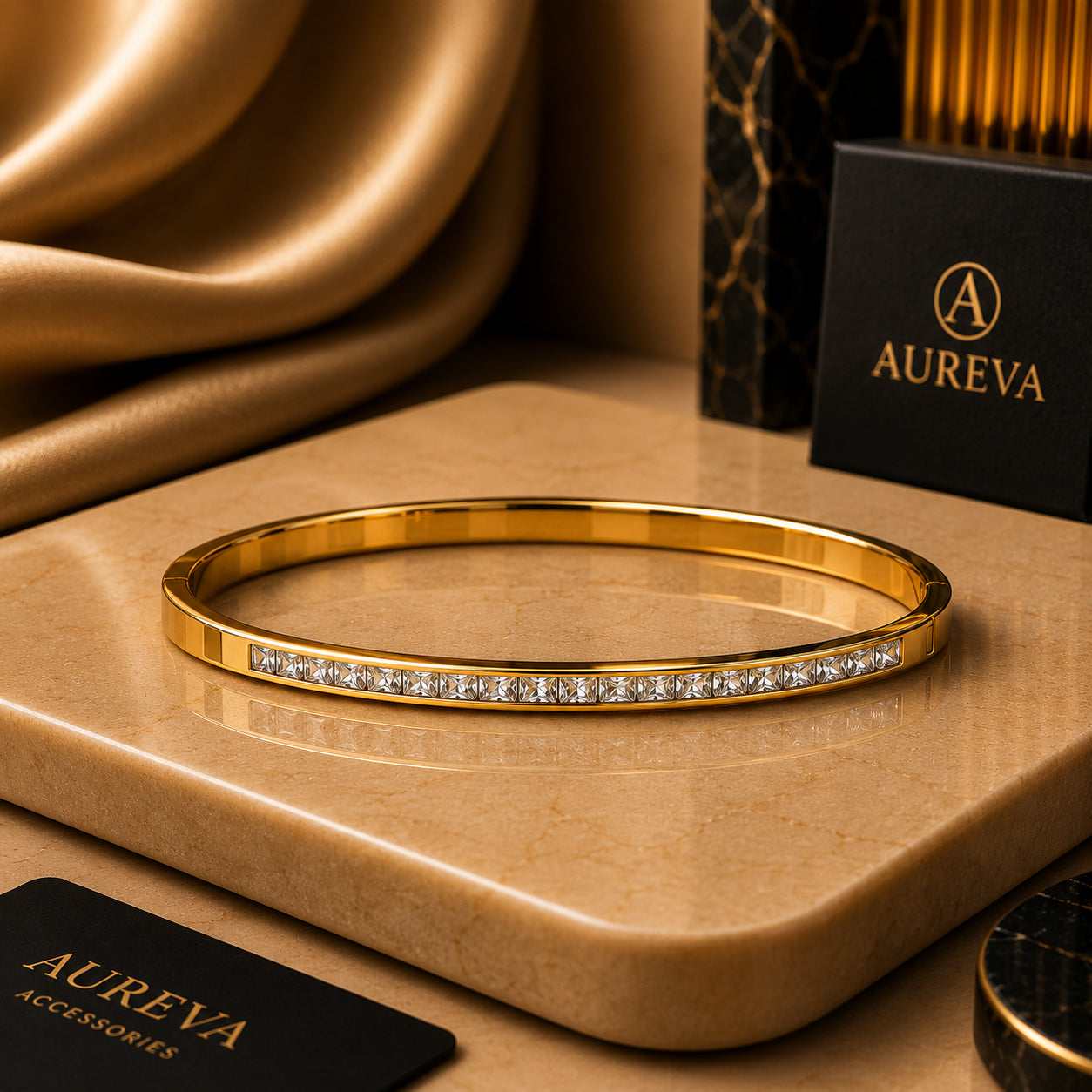 A gold bangle bracelet with a row of clear stones on the front, displayed on a beige stone slab with gold fabric and an Aureva box in the background.