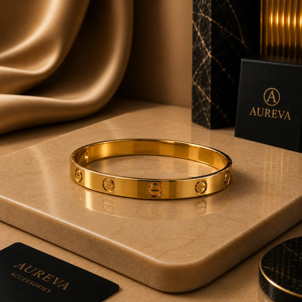 A polished gold bangle bracelet with screw details, displayed on a beige marble slab with luxury packaging and draped gold fabric in the background. The brand name AUREVA is visible on the packaging.