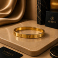 A polished gold bangle bracelet with screw details, displayed on a beige marble slab with luxury packaging and draped gold fabric in the background. The brand name AUREVA is visible on the packaging.
