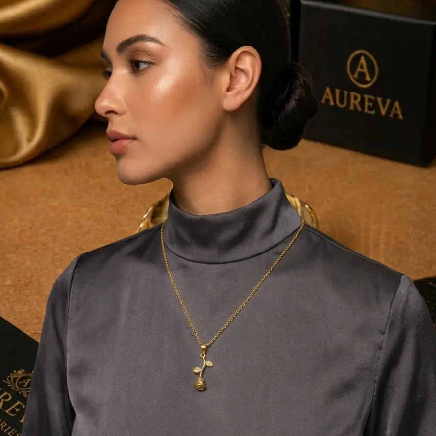 A woman wearing a gold cross pendant necklace with a small rose gold heart detail, styled with a dark gray mock neck top. Aureva Accessories branding is visible on the background boxes.