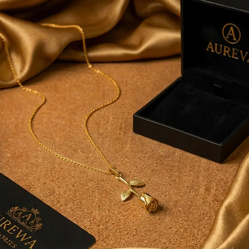 A flat gold rose pendant necklace resting on a beige cork surface next to a black Aureva jewelry box and draped gold silk fabric.