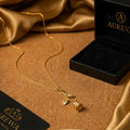 A flat gold rose pendant necklace resting on a beige cork surface next to a black Aureva jewelry box and draped gold silk fabric.