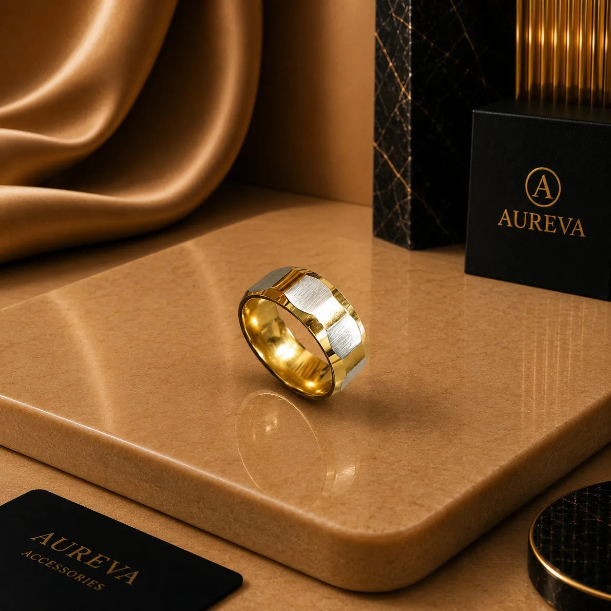 A gold and silver band ring displayed on a polished tan surface, with Aureva branding visible on a black box and card in the scene.