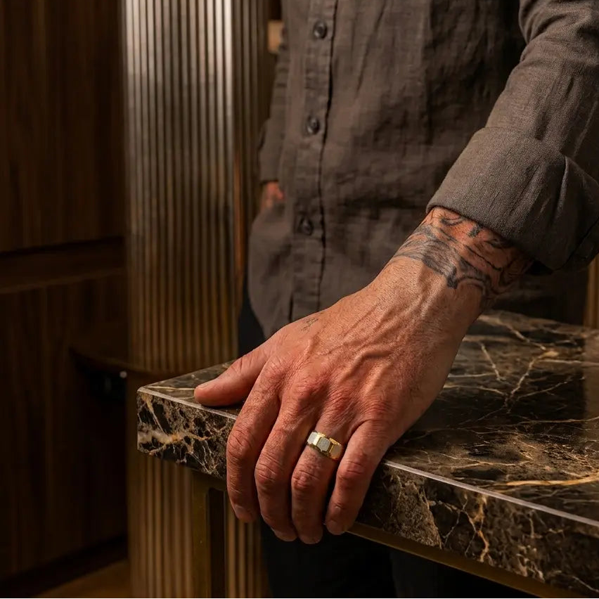 A close-up of a man's hand resting on a dark marble countertop, wearing a silver band ring. He is wearing a dark brown linen button-up shirt, with a modern ribbed wood-paneled interior in the background.