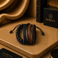 A set of four men's leather and bead bracelets displayed on a textured brown surface with gold satin fabric and branded black packaging in the background. The bracelets include black beaded and braided styles with brown and black leather bands.
