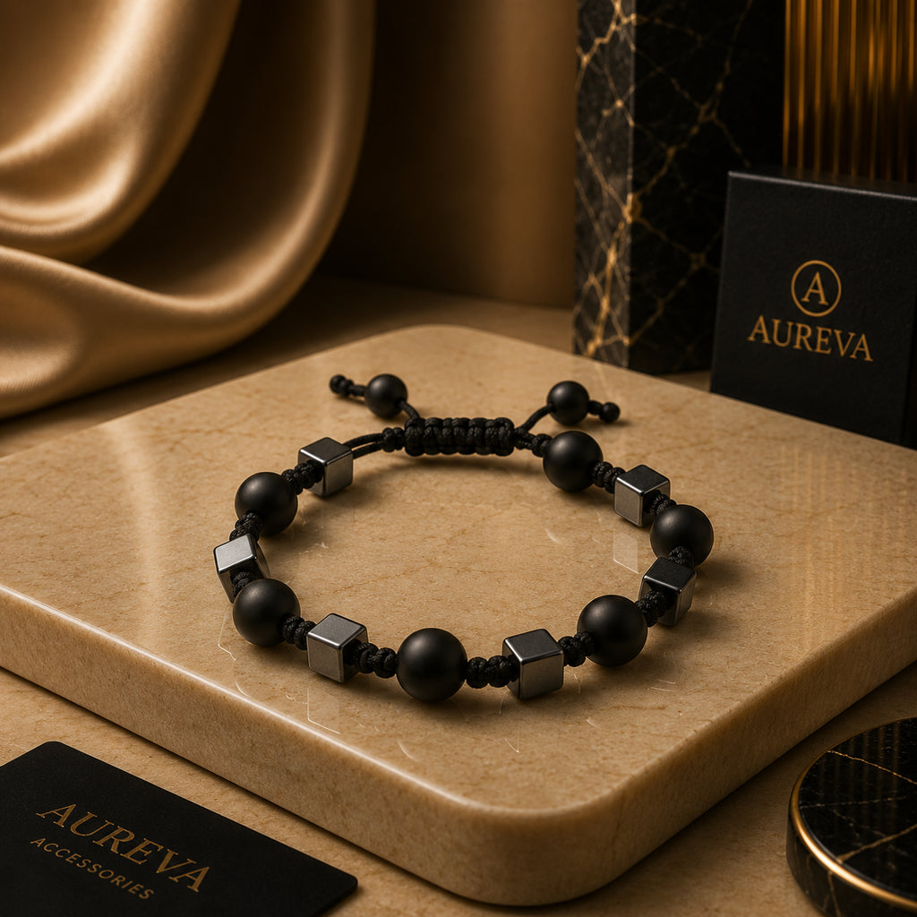 A black beaded bracelet with matte black round beads and metallic silver cube accents, displayed on a beige stone slab with Aureva accessories branding in the background.