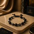 A black beaded bracelet with matte black round beads and metallic silver cube accents, displayed on a beige stone slab with Aureva accessories branding in the background.