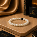 A beaded bracelet with mostly white round beads and one black accent bead, displayed on a tan stone slab. Black Aureva accessories boxes are visible in the background and foreground, with warm draped fabric and a dark marble-textured object behind.