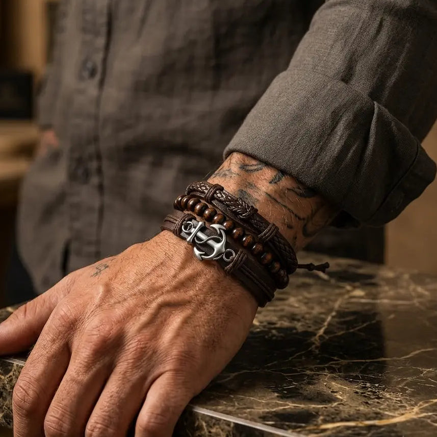 A close-up of a man's tattooed wrist wearing a stack of dark brown leather bracelets, including one with a silver skull-and-crossbones charm, resting on a dark marble tabletop.