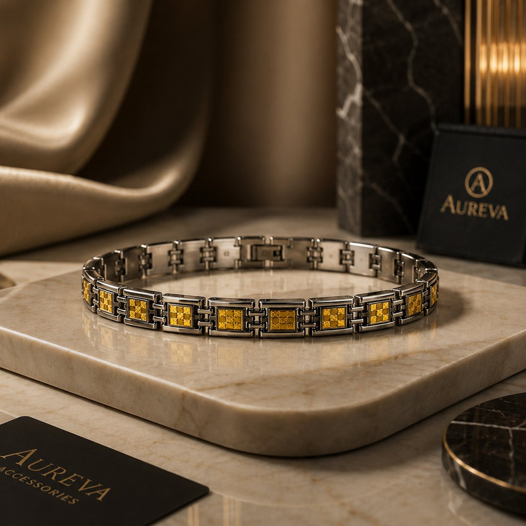 A silver-toned metal bracelet with repeating square links featuring yellow-gold patterned inlays, displayed on a beige marble slab. Aureva brand packaging and a draped satin fabric are visible in the background.