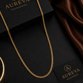 A gold chain necklace draped elegantly over a black presentation box and card, with a brown satin fabric in the background. The brand name 'AUREVA ACCESSORIES' is clearly visible on the packaging.