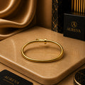 A polished gold bangle bracelet displayed on a beige stone slab, with draped gold fabric in the background and an Aureva Accessories box visible.