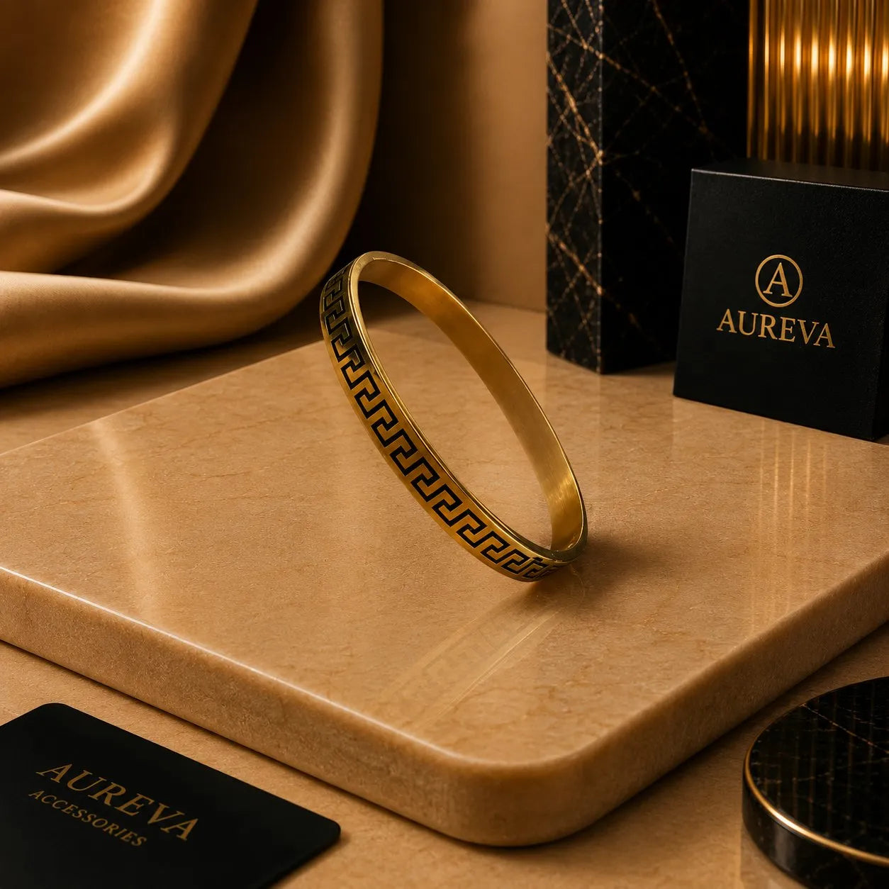 A gold bangle bracelet with a black Greek key pattern, displayed on a beige stone slab with luxury brand packaging in the background.