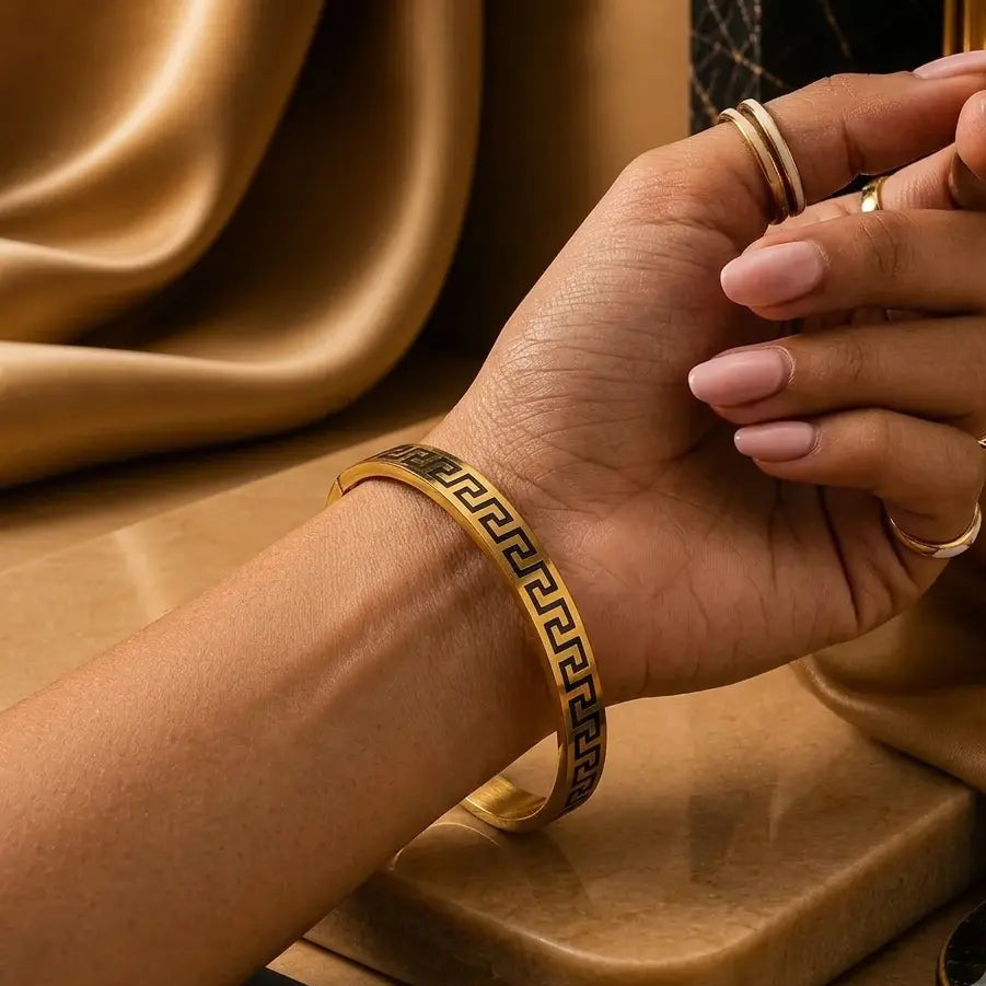 A gold-toned bangle bracelet with a black Greek key pattern, worn on a woman's wrist. The hand has light pink manicured nails and is adorned with gold rings, set against a luxurious background of draped gold satin fabric and a polished stone slab.