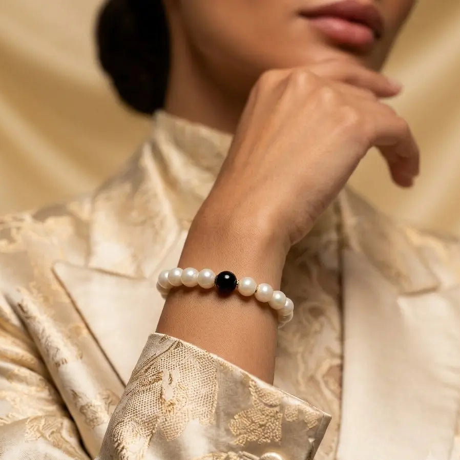 A close-up of a woman wearing a pearl beaded bracelet with a single black bead at the center, styled with a gold-patterned satin jacket against a warm beige background.